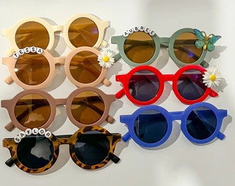 Può includere: Set di otto occhiali da sole rotondi per bambini, con vari colori e design. Alcuni occhiali da sole hanno un abbellimento a margherita o farfalla, mentre altri hanno nomi scritti in perline.