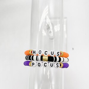 May include: Three bracelets with the word "HOCUS POCUS" spelled out in black letters on white beads. The bracelets are layered on top of each other, with the top bracelet being orange, the middle bracelet being black and white, and the bottom bracelet being purple.