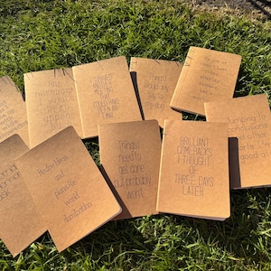 May include: A selection of kraft paper notebooks, each with a different humorous quote printed in black. The notebooks are displayed on green grass. Quotes include "My notebook I write in to appear busy."