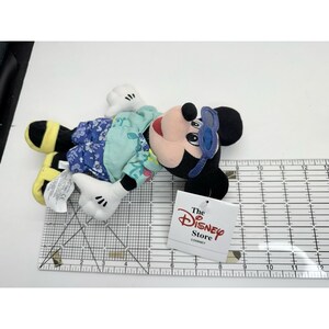 May include: A Mickey Mouse plush toy with sunglasses, wearing a floral patterned shirt. The toy has black ears, a red mouth, and white gloves. A tag from The Disney Store is attached. The toy measures approximately 12 inches in length.