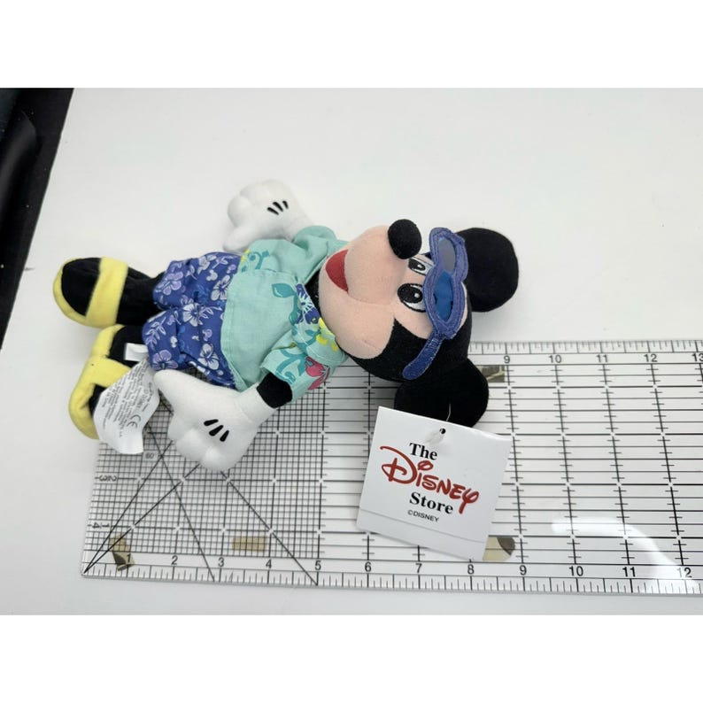May include: A Mickey Mouse plush toy with sunglasses, wearing a floral patterned shirt. The toy has black ears, a red mouth, and white gloves. A tag from The Disney Store is attached. The toy measures approximately 12 inches in length.
