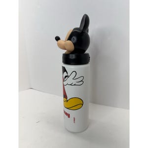 May include: White water bottle featuring a Mickey Mouse design. The bottle has a black lid and a Mickey Mouse head-shaped top. The design includes Mickey's face, arms, and feet in red, yellow, and black. The word "hey" is printed on the bottle.