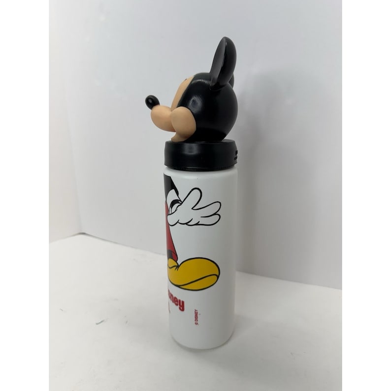 May include: White water bottle featuring a Mickey Mouse design. The bottle has a black lid and a Mickey Mouse head-shaped top. The design includes Mickey's face, arms, and feet in red, yellow, and black. The word "hey" is printed on the bottle.