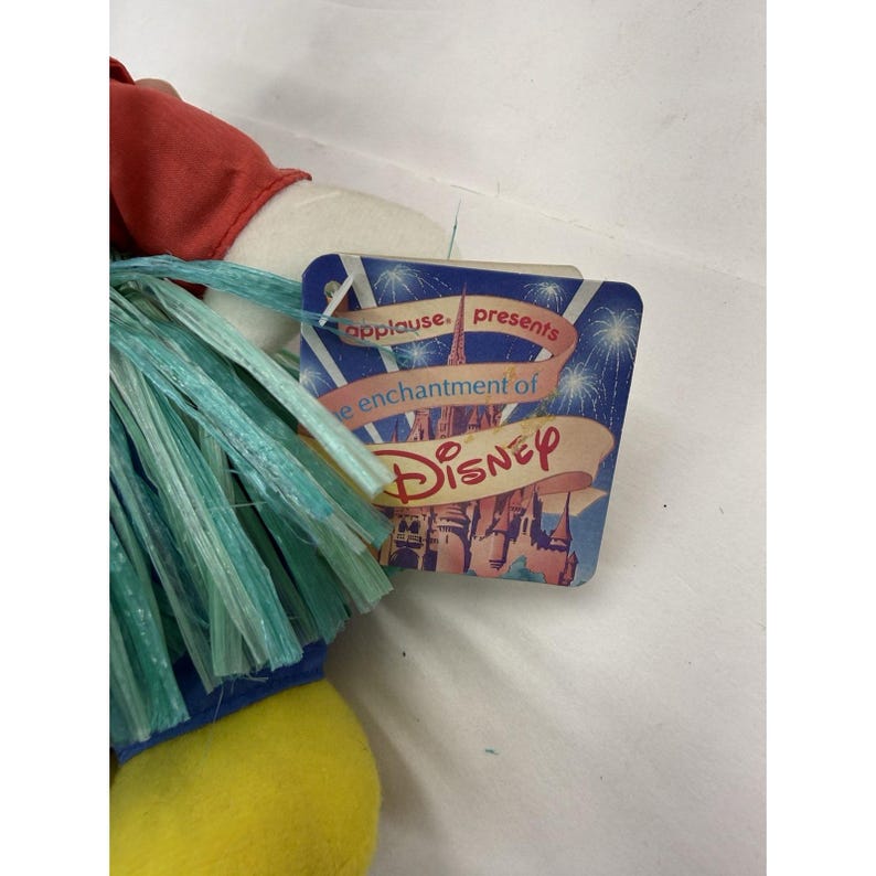 Puede incluir: Un peluche de un personaje de Disney con una parte superior roja, detalles azules y una parte inferior amarilla. El personaje tiene flecos azules y verdes y una etiqueta que dice "applause presents the enchantment of Disney."