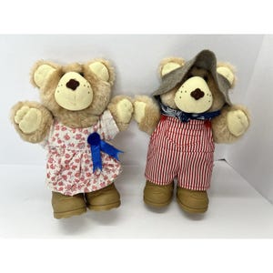May include: Two plush teddy bears. One bear wears a floral dress with a blue bow, the other wears red and white striped overalls, a hat, and a blue bandana. Both bears have tan fur and brown noses.