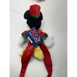 Puede incluir: Un peluche de Mickey Mouse con un atuendo rojo y verde y un sombrero a juego. El juguete tiene guantes blancos y zapatos amarillos. Una etiqueta dice "100 Years of Magic" con gr&aacute;ficos de Mickey Mouse.