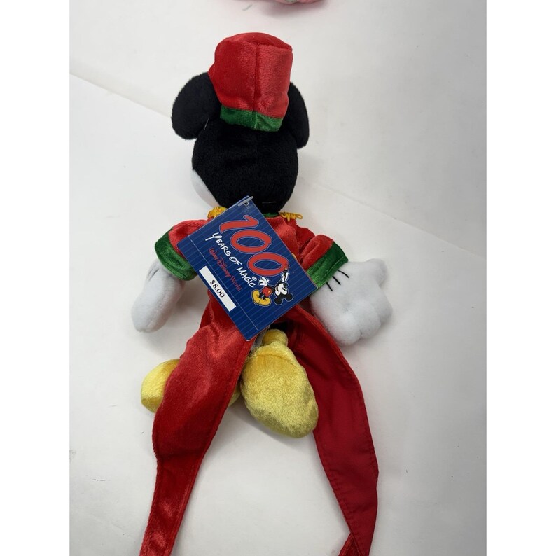 Puede incluir: Un peluche de Mickey Mouse con un atuendo rojo y verde y un sombrero a juego. El juguete tiene guantes blancos y zapatos amarillos. Una etiqueta dice "100 Years of Magic" con gr&aacute;ficos de Mickey Mouse.