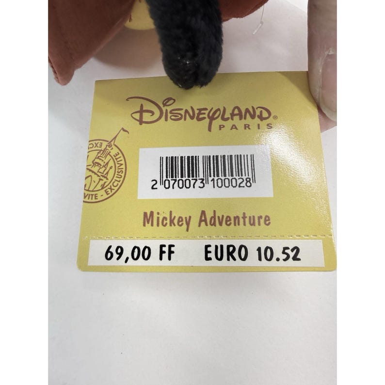 K&ouml;nnte beinhalten: Ein Disneyland Paris-Etikett mit dem Text "Mickey Adventure" und einem Barcode. Das Etikett ist gelb und hat oben das Disneyland Paris-Logo. Das Etikett ist an einem Pl&uuml;schtier befestigt.