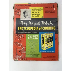 May include: A vintage cookbook titled "Mary Margaret McBride Encyclopedia of Cooking." The cover is red, black, and white, with text and illustrations. It features a portrait of a woman and promises exciting dishes from around the world.