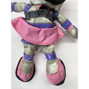 May include: A plush toy with a silver and pink outfit. The toy has a purple belt and purple accents on the shoes. The toy is wearing a pink skirt.