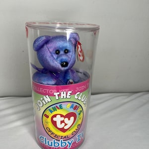 May include: A purple and blue plush bear toy inside a clear plastic container. The container has a colorful label with the text "Join the Club" and "Beanie Babies Official Club". The bear has a red tag attached.
