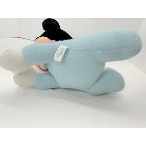 Puede incluir: Un peluche de Mickey Mouse tumbado boca arriba. El juguete tiene una cabeza negra, un cuerpo azul claro y manos y pies blancos. Una etiqueta est&aacute; adherida al cuerpo del juguete. El juguete es un producto de Disney.