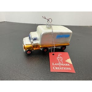 May include: A glass ornament shaped like a white delivery truck with gold trim and red wheels. The side of the truck features the blue text UNITED. A silver hanging loop is attached to the top, and a red tag reads LANDMARK CREATIONS.