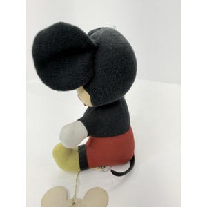 Puede incluir: Un mu&ntilde;eco de peluche vintage de Mickey Mouse. El mu&ntilde;eco tiene cabeza y orejas negras, cuerpo negro, pantalones cortos rojos, pies amarillos y guantes blancos. El mu&ntilde;eco est&aacute; sentado y tiene una etiqueta.