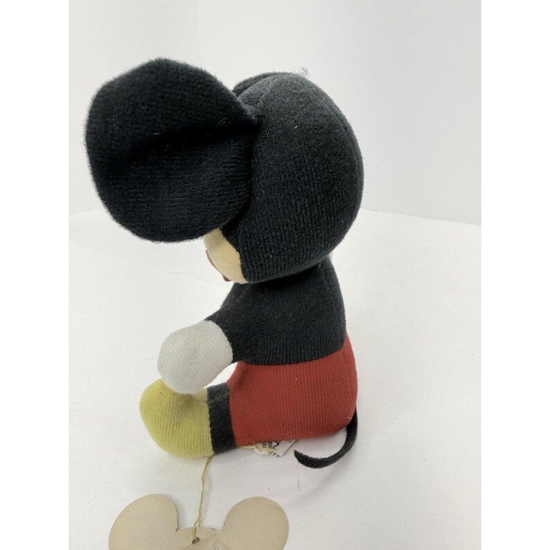 Puede incluir: Un mu&ntilde;eco de peluche vintage de Mickey Mouse. El mu&ntilde;eco tiene cabeza y orejas negras, cuerpo negro, pantalones cortos rojos, pies amarillos y guantes blancos. El mu&ntilde;eco est&aacute; sentado y tiene una etiqueta.