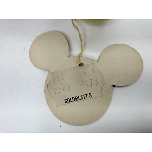 Puede incluir: Una etiqueta vintage con forma de Mickey Mouse en beige. La etiqueta tiene una etiqueta de precio rectangular con el texto "GOLDBLATT'S" y n&uacute;meros. Una cuerda amarilla est&aacute; unida a la parte superior. La etiqueta est&aacute; sobre una superficie blanca.