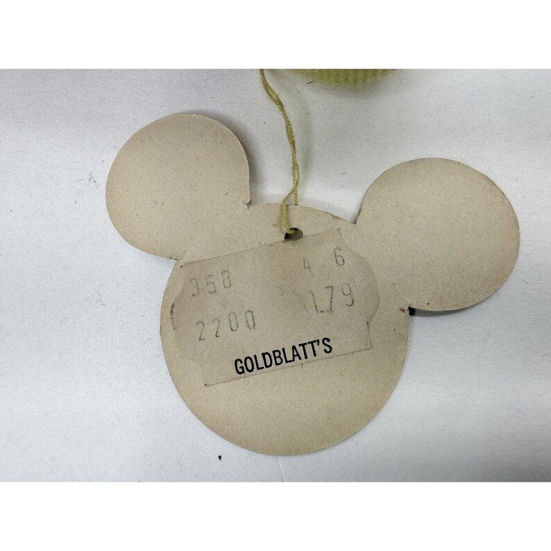 Puede incluir: Una etiqueta vintage con forma de Mickey Mouse en beige. La etiqueta tiene una etiqueta de precio rectangular con el texto "GOLDBLATT'S" y n&uacute;meros. Una cuerda amarilla est&aacute; unida a la parte superior. La etiqueta est&aacute; sobre una superficie blanca.