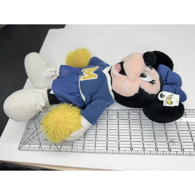 May include: A plush Minnie Mouse doll wearing a blue and gold cheerleader outfit. The doll has black ears, a pink face, and white gloves and shoes. The doll is lying down on a white surface.