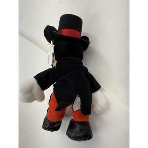 Peut inclure: Peluche Mickey Mouse, vue de dos, portant un chapeau haut de forme noir avec un ruban rouge, une veste noire, un pantalon rouge et des chaussures noires. Il a des gants blancs et une &eacute;tiquette.