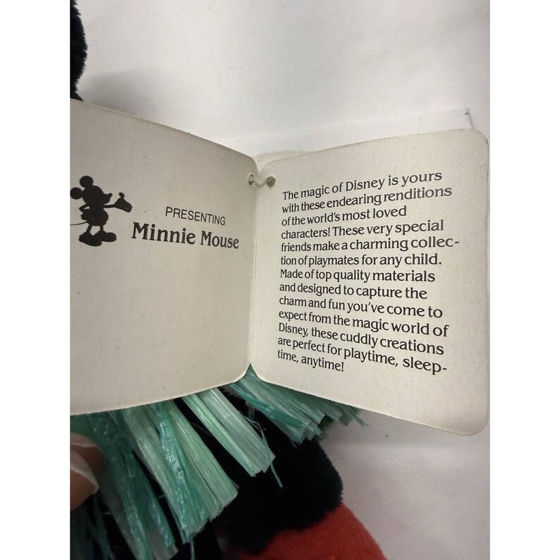 Puede incluir: Un peluche de Minnie Mouse con un lazo rojo y pelaje negro. La etiqueta dice "PRESENTING Minnie Mouse" con un logotipo de Disney. La etiqueta tambi&eacute;n incluye texto sobre el juguete.