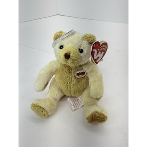 May include: A plush teddy bear with a cream-colored head and limbs, and a tan body. The bear has a silver band around its head and a red heart-shaped tag. The bear is approximately 8 inches tall.