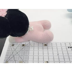 Puede incluir: Un juguete de peluche rosa con pies blancos y una cabeza negra. El juguete tiene un lazo blanco y est&aacute; sobre una superficie blanca junto a una regla.