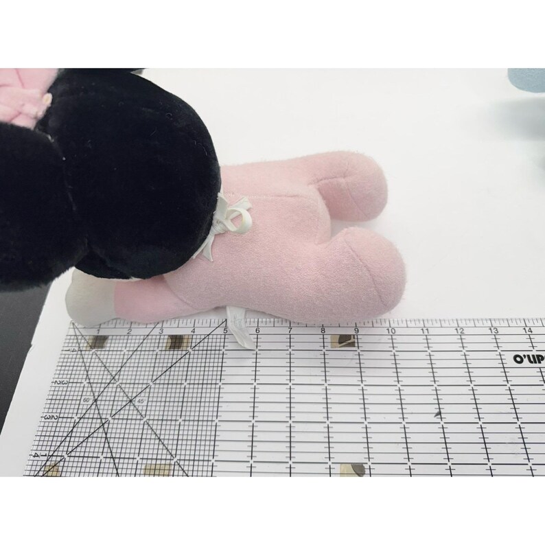Puede incluir: Un juguete de peluche rosa con pies blancos y una cabeza negra. El juguete tiene un lazo blanco y est&aacute; sobre una superficie blanca junto a una regla.