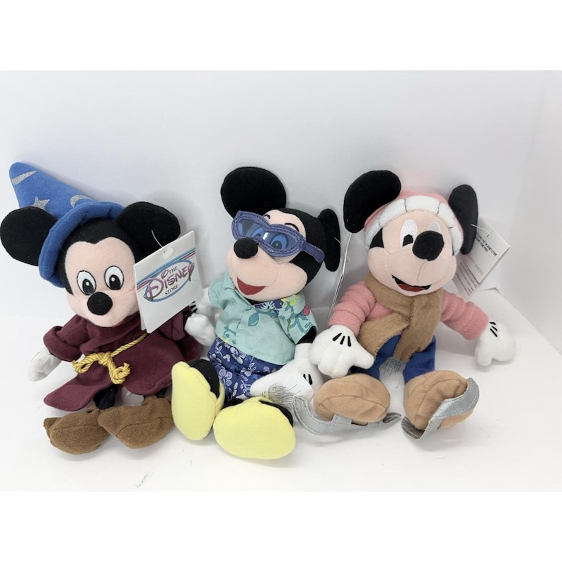 May include: Three Mickey Mouse plush toys in different outfits. One wears a sorcerer's hat and robe, another has sunglasses and a Hawaiian shirt, and the third is dressed for winter with a hat and scarf. Each toy has a Disney Store tag.