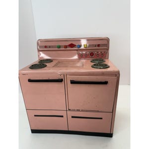 May include: A vintage pink toy stove with four burners, two oven doors, and a top panel with colorful knobs and a diamond-shaped emblem. The stove is made of metal and has black accents.