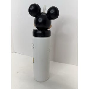 May include: White water bottle featuring a Mickey Mouse design. The bottle has a black lid with a Mickey Mouse head and ears on top, and a white straw. The bottle has a white body with a few Mickey Mouse details.
