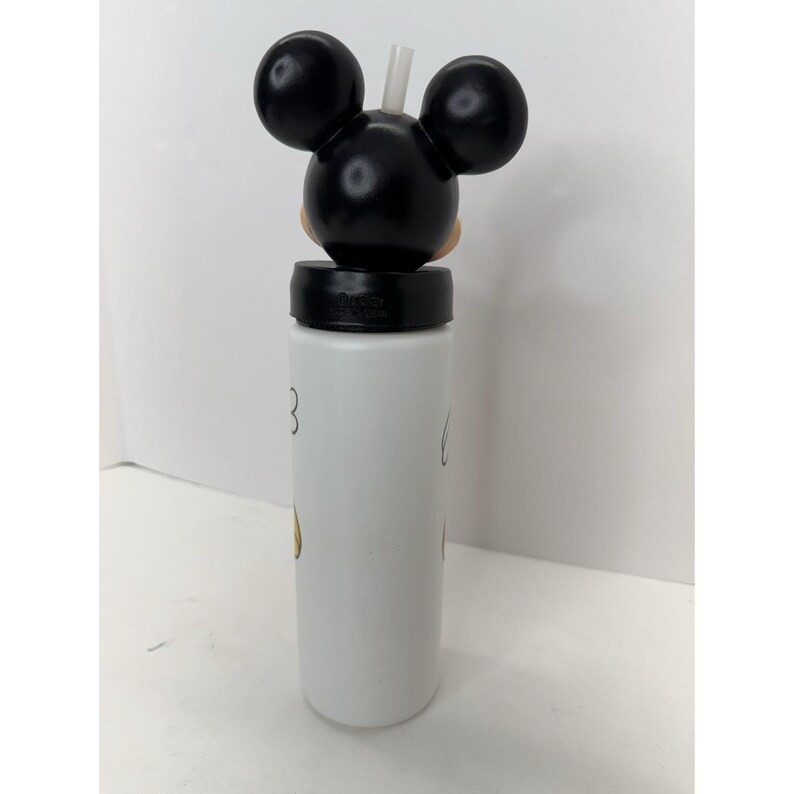 May include: White water bottle featuring a Mickey Mouse design. The bottle has a black lid with a Mickey Mouse head and ears on top, and a white straw. The bottle has a white body with a few Mickey Mouse details.