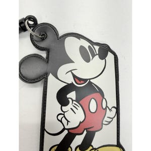 May include: A black and white Mickey Mouse luggage tag with a black border and a black strap. The tag features Mickey Mouse in his classic outfit, with red shorts, white gloves, and yellow shoes. The tag is rectangular with a Mickey Mouse head shape at the top.