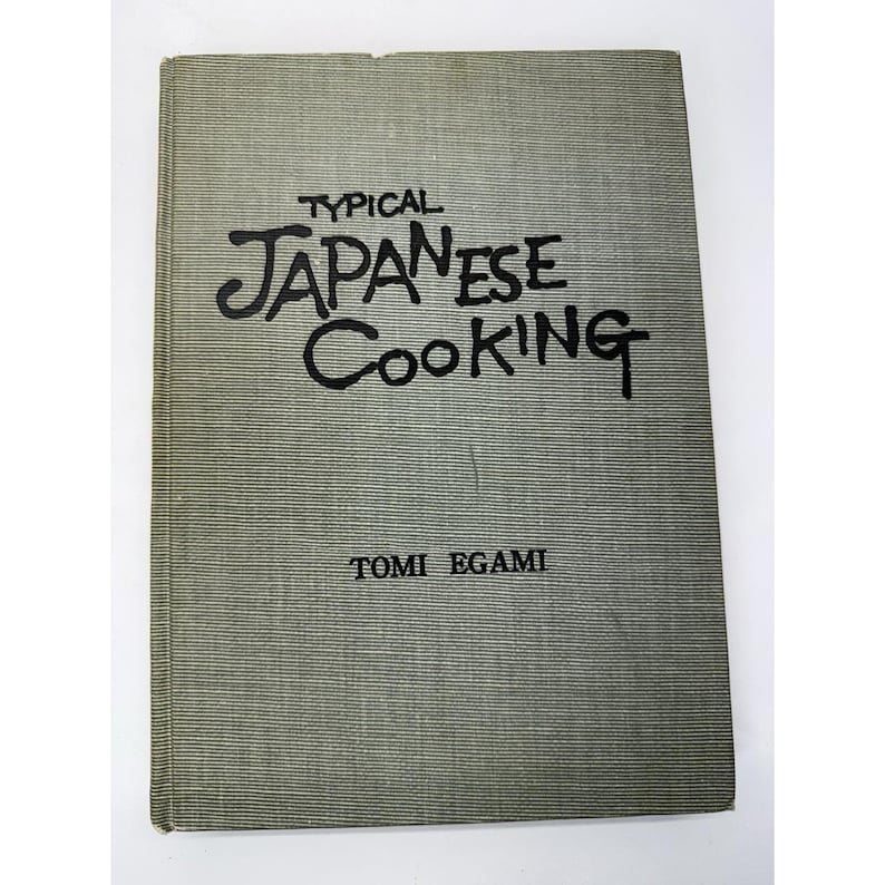 May include: A vintage cookbook titled "Typical Japanese Cooking" by Tomi Egami. The book has a textured, gray cover with the title and author's name in black lettering. The cover is slightly worn, suggesting its age.