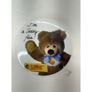 May include: A round white button featuring a brown teddy bear with a blue bow tie. The text "I'm a Teddy fan!" is written above the bear. The button also has the Steiff logo and the words "BUTTON IN EAR".