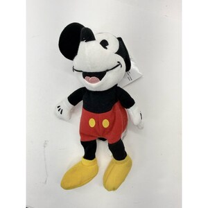 Puede incluir: Un peluche de Mickey Mouse. El juguete tiene una cara blanca, orejas negras y un cuerpo negro. Lleva pantalones cortos rojos con botones amarillos y zapatos amarillos. El juguete est&aacute; sonriendo y tiene una lengua rosa.