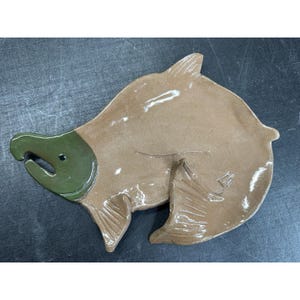 May include: A ceramic dish shaped like a fish with a glossy finish. The head is dark olive green with a small black eye, while the body and fins are a light tan brown color. The surface features incised lines to define the fins and gills.