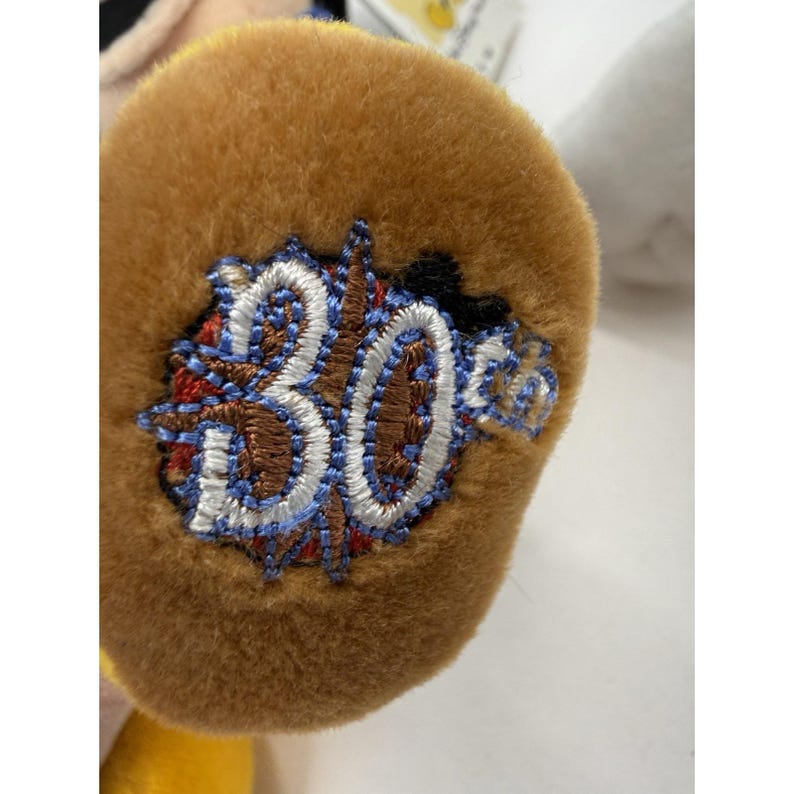 Peut inclure: Gros plan sur la patte marron d'une peluche avec un &eacute;cusson brod&eacute;. L'&eacute;cusson porte le chiffre "30" en points blancs, entour&eacute; d'un motif bleu, rouge et marron. La peluche est jaune et blanche.