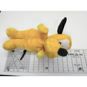 Puede incluir: Un peluche de Pluto, un perro amarillo con orejas y nariz negras, y un collar rojo. El juguete est&aacute; tumbado boca arriba. El juguete est&aacute; sobre una superficie blanca. El juguete mide unos 25 cm de largo.
