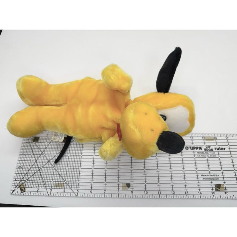 Puede incluir: Un peluche de Pluto, un perro amarillo con orejas y nariz negras, y un collar rojo. El juguete est&aacute; tumbado boca arriba. El juguete est&aacute; sobre una superficie blanca. El juguete mide unos 25 cm de largo.
