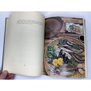 May include: An open cookbook page with a food photography spread. The right page features a woven basket filled with seafood, vegetables, and flowers. A small dish of dipping sauce and a pan are also visible. The left page has text.