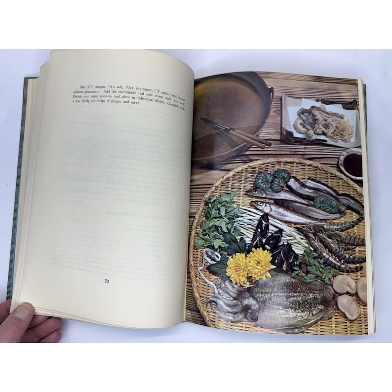 May include: An open cookbook page with a food photography spread. The right page features a woven basket filled with seafood, vegetables, and flowers. A small dish of dipping sauce and a pan are also visible. The left page has text.