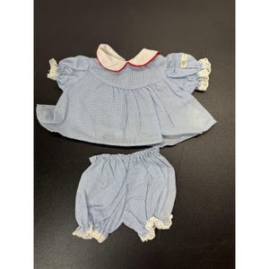 May include: A light blue and white gingham top and matching bloomers set. The top features a white Peter Pan collar with red piping, short puffed sleeves with white eyelet lace trim, and a gathered bodice. The bloomers have white eyelet lace trim.