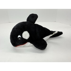 May include: A plush orca whale toy. The toy is primarily black with white patches around the eyes and on the sides. It has a red mouth and a small white tag on the tail. The orca has a friendly, cartoonish appearance.