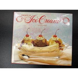 May include: A hardcover book titled The Joy of Ice Cream by Matthew Klein. The cover features a banana split with three scoops of ice cream topped with whipped cream and cherries in a glass dish, with a silver spoon resting on a cloth napkin.