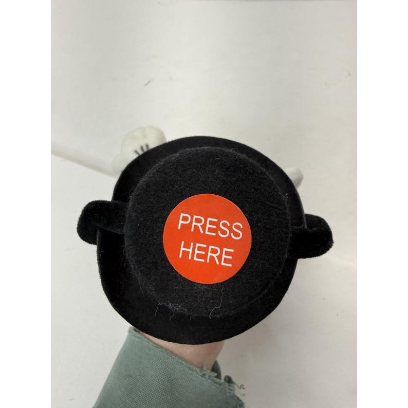 Peut inclure: Un chapeau en peluche noir avec un autocollant rond rouge qui dit "PRESS HERE" en texte blanc. Le chapeau a deux petites protub&eacute;rances en forme d'oreille de chaque c&ocirc;t&eacute;. Le chapeau est tenu par une personne portant une chemise verte.