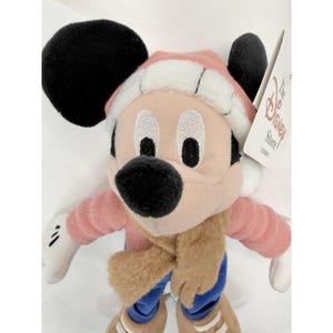 May include: A plush Mickey Mouse doll wearing a pink and white winter hat, pink long-sleeve shirt, brown scarf, blue pants, and brown shoes. The doll has black ears and a black nose. A tag from The Disney Store is attached.