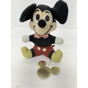 Puede incluir: Un peluche vintage de Mickey Mouse, con cuerpo, orejas y nariz negros, cara color crema y manos blancas. El peluche lleva pantalones cortos rojos, pies amarillos y una pajarita rosa. Una etiqueta dorada con "Mickey Mouse" est&aacute; adjunta.