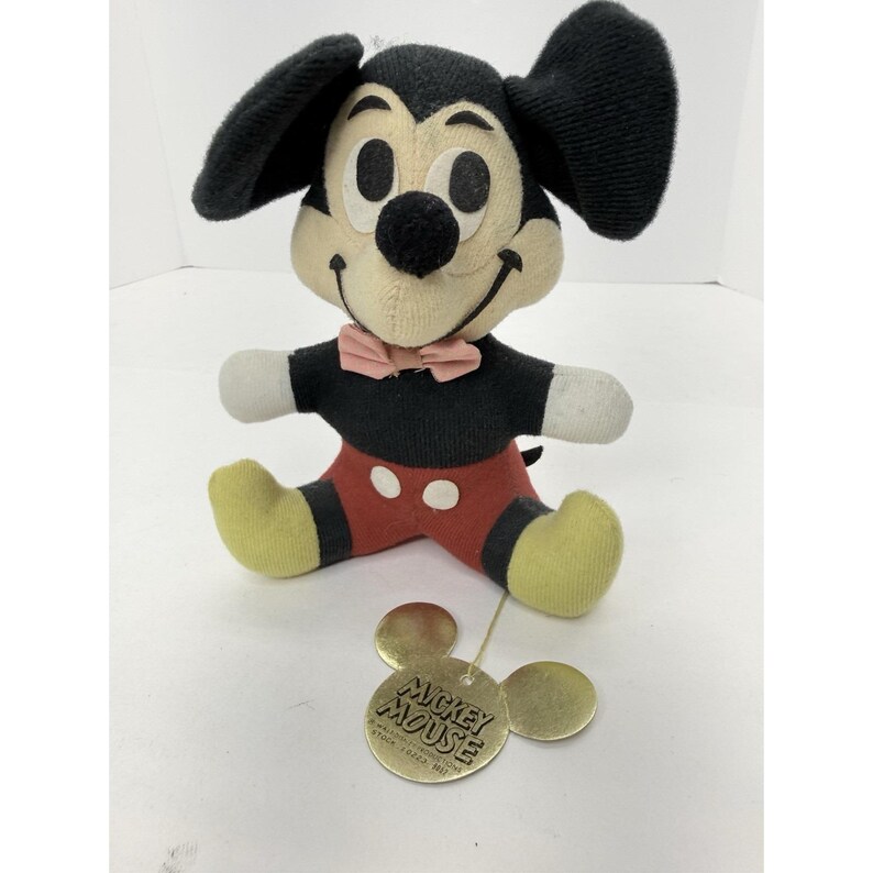 Puede incluir: Un peluche vintage de Mickey Mouse, con cuerpo, orejas y nariz negros, cara color crema y manos blancas. El peluche lleva pantalones cortos rojos, pies amarillos y una pajarita rosa. Una etiqueta dorada con "Mickey Mouse" est&aacute; adjunta.