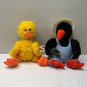 May include: Two plush toys, a yellow chick and a black bird wearing overalls and a straw hat. The chick has a red beak and orange feet. The bird has a red beak and orange feet. Both toys have Ty tags.