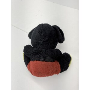 May include: A plush toy of a cartoon character, viewed from the back. The toy is primarily black with large, rounded ears. It has a red lower body and yellow accents on the feet. The toy is sitting on a white surface.